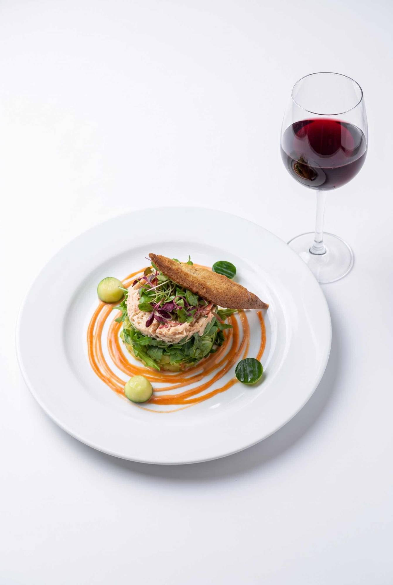 Dressed crab on avocado and rocket with a Melba toast, cucumber and a Marie Rose swirl, beside a glass of red wine